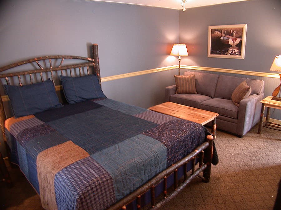 A cozy bedroom with a rustic log-style queen bed featuring a blue patchwork quilt, a small sofa, and blue and yellow walls.