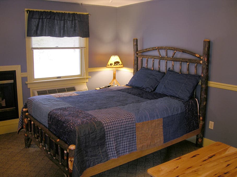 A cozy bedroom featuring a rustic log-style bed with a blue patchwork quilt, a window with a coordinating valance, and a small fireplace alcove.