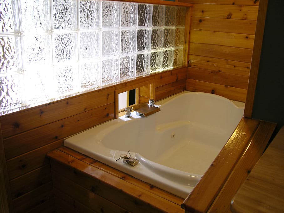 A close-up of a white jetted tub set in a wood-paneled alcove with natural pine walls and a long panel of glass blocks for light.