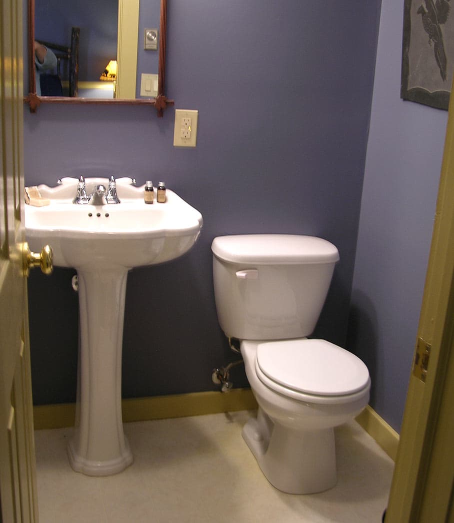 A small, bright bathroom with lavender blue walls, featuring a white pedestal sink and a white toilet, with a wooden-framed mirror visible.