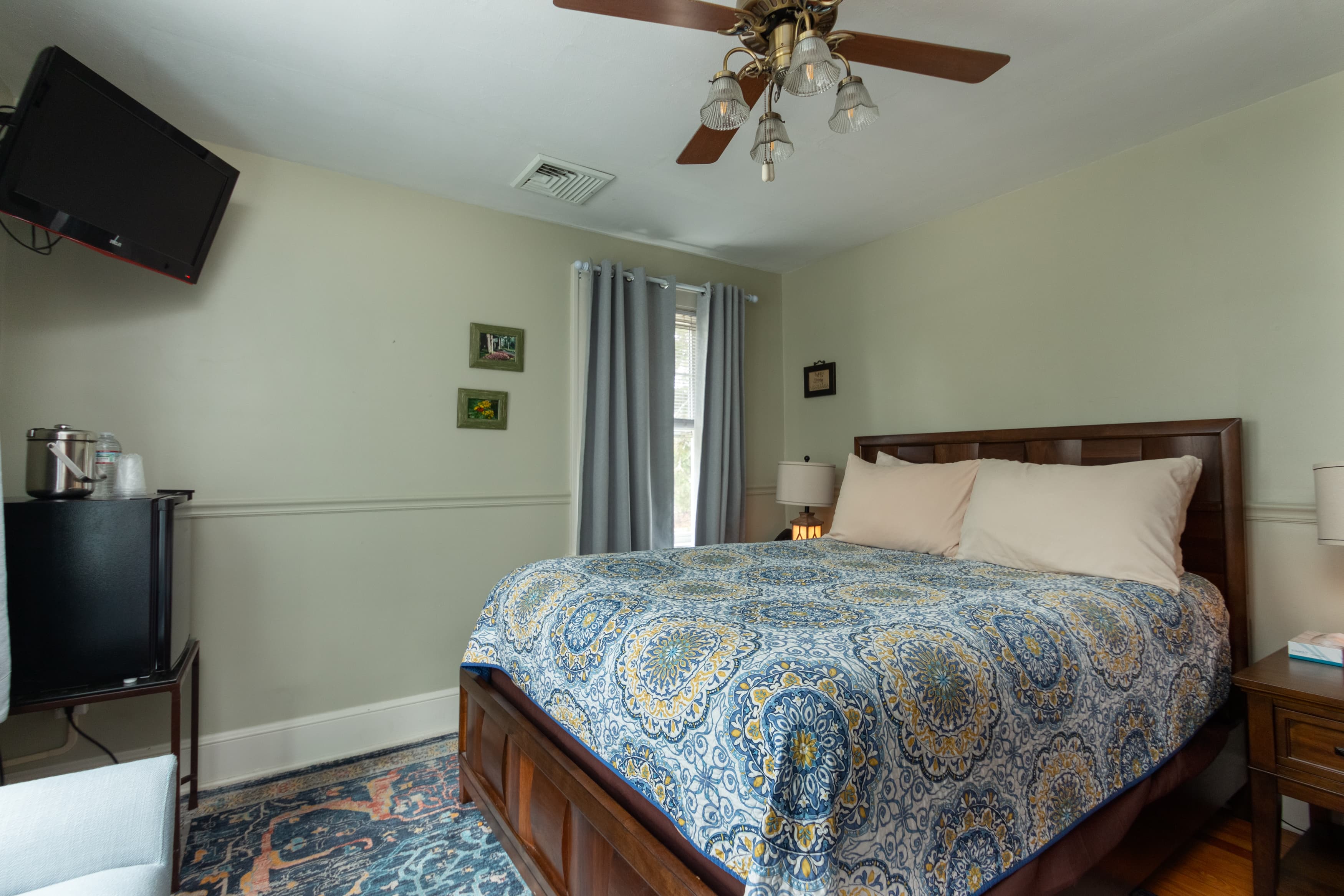 A bright bedroom with light green walls, a queen bed featuring a blue and yellow medallion quilt, a wall-mounted TV, and a ceiling fan.