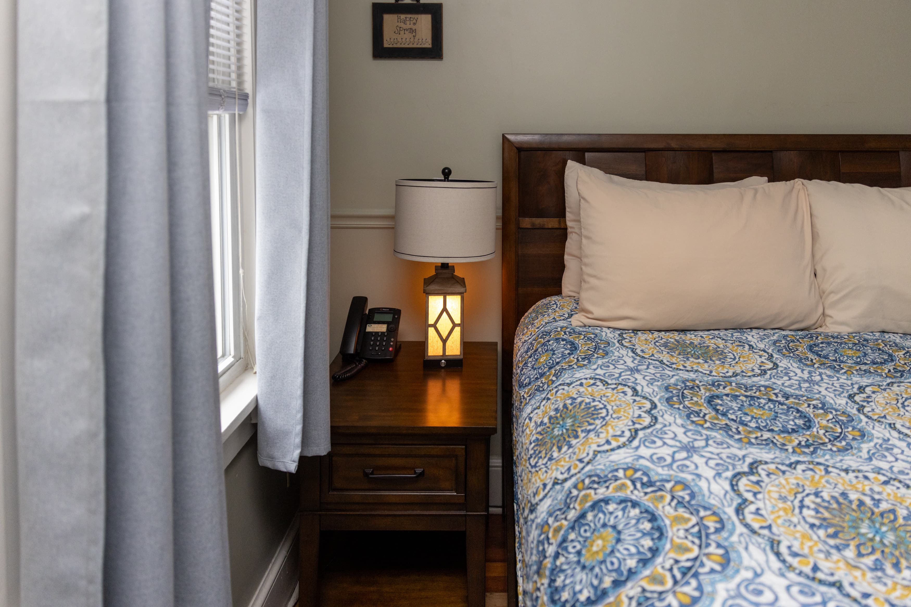 A close-up of a dark wood nightstand next to a queen bed with a blue and yellow quilt, featuring a lantern-style lamp and a phone.