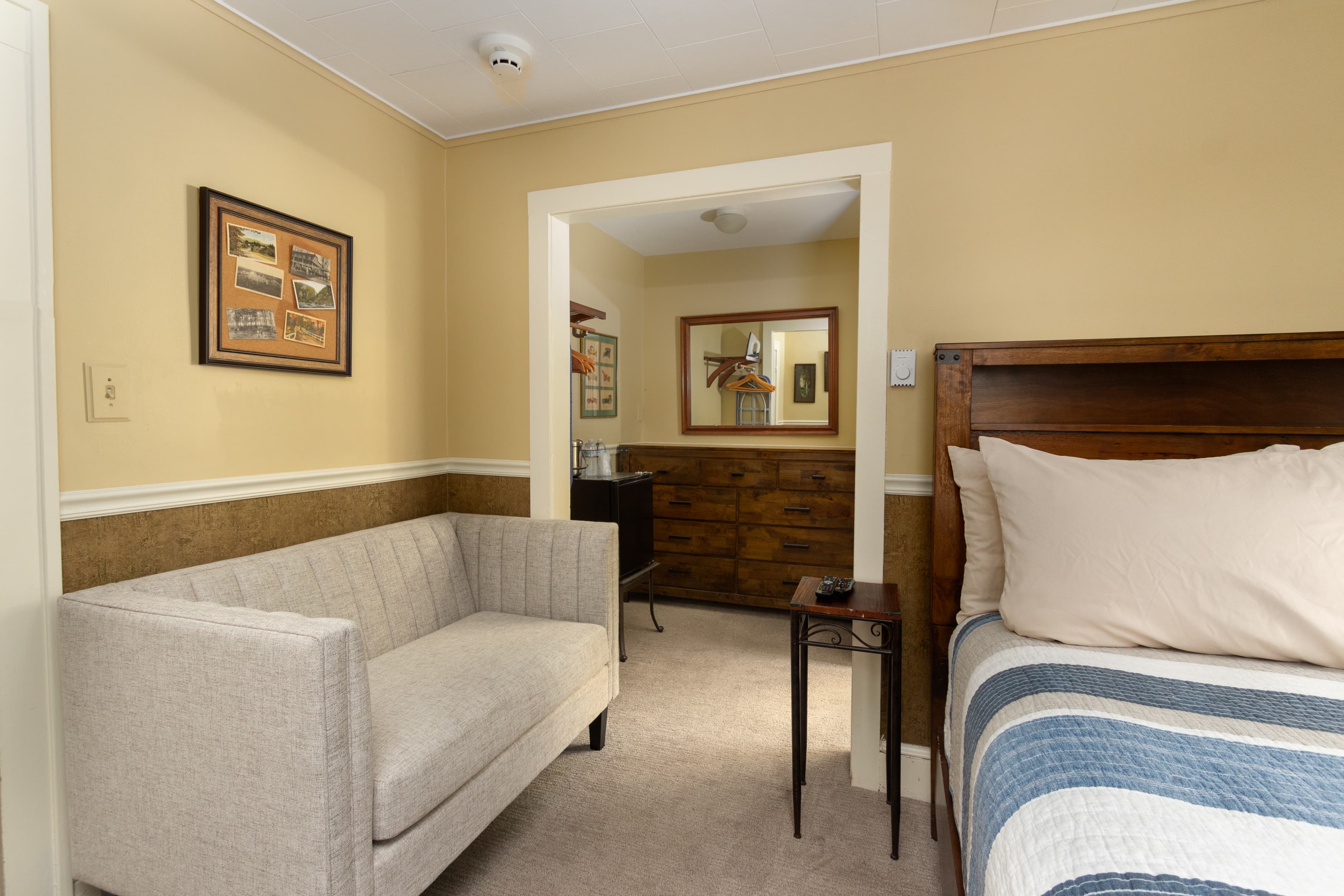 A suite with a light-colored settee in the foreground and a dark wood bed partially visible, looking into a dressing area.