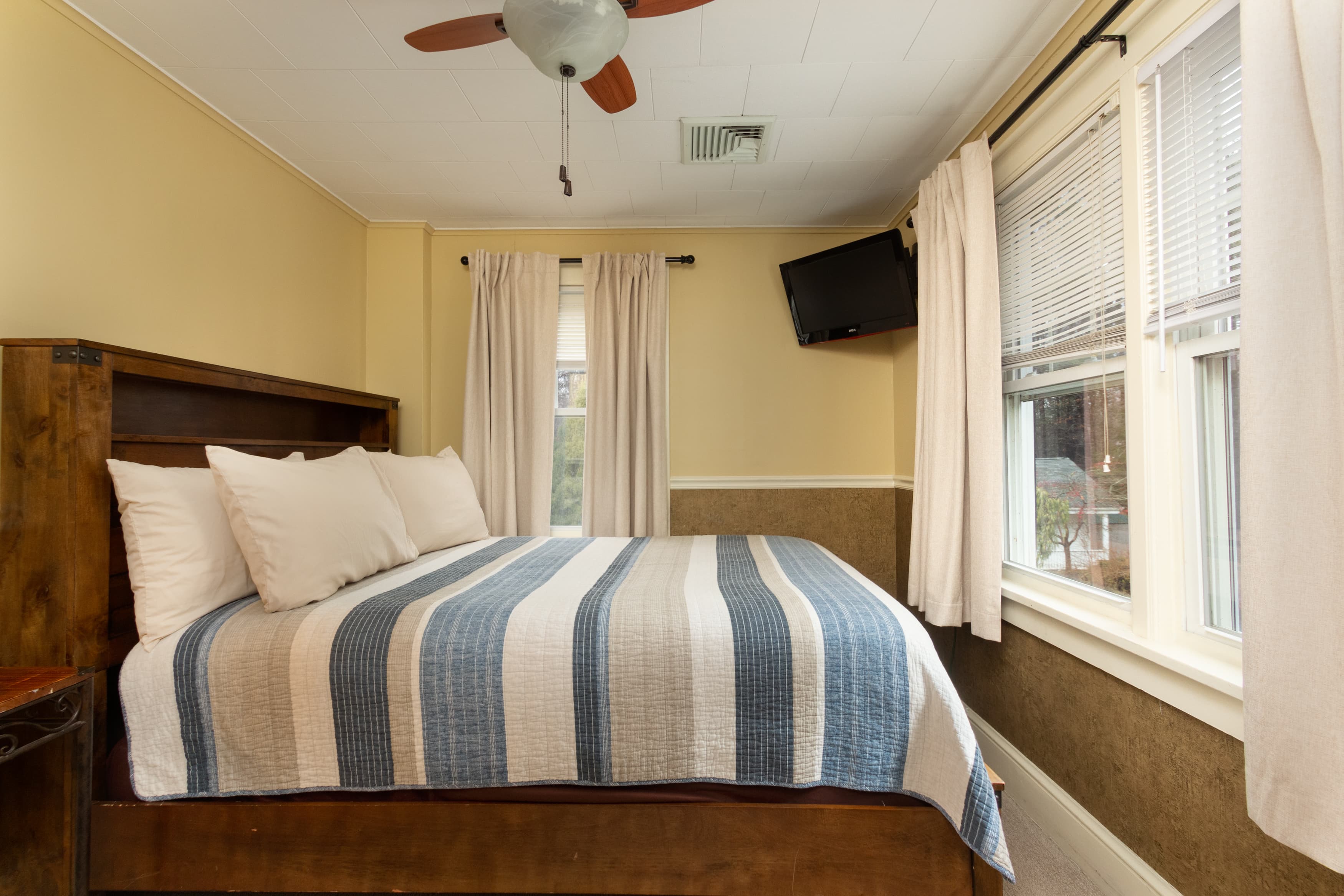A cozy bedroom with a dark wood bed frame, a blue and white striped quilt, pale yellow walls, and a wall-mounted TV.