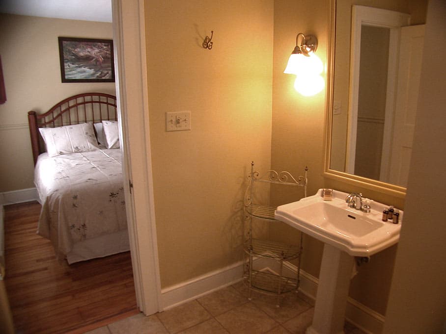 A small vanity area with a white pedestal sink and sconce light, looking through an open doorway into a cozy bedroom with a wood headboard.