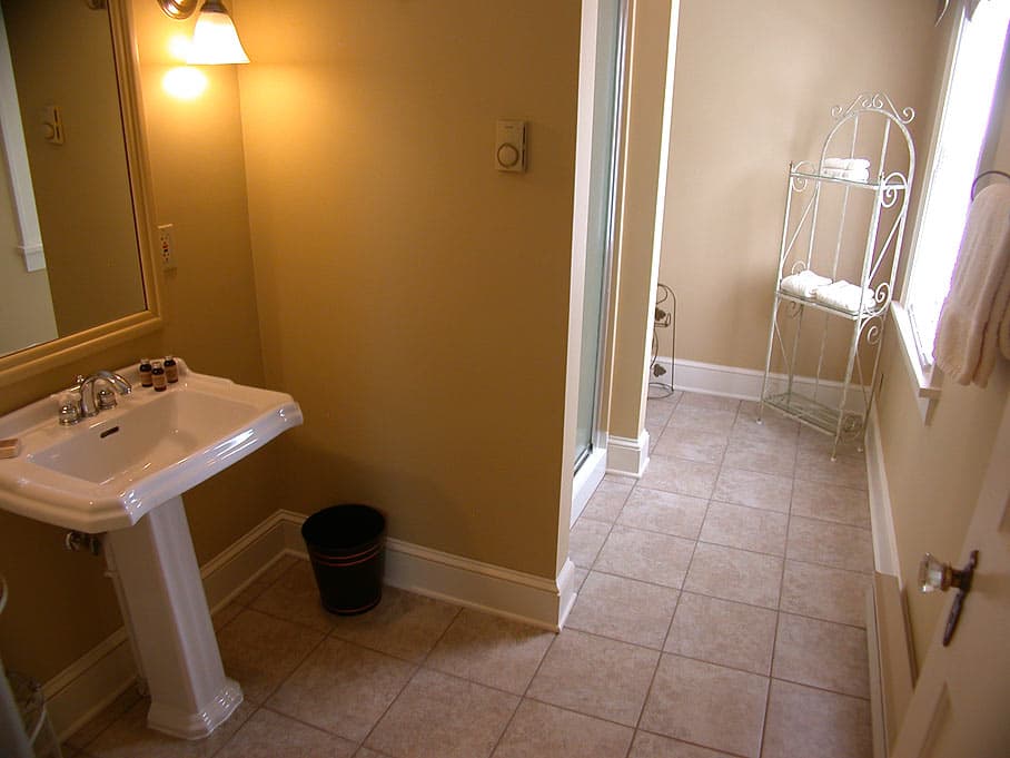 A simple, spacious bathroom with tan walls, a white pedestal sink, and tiled floors leading to a shower enclosure and window.