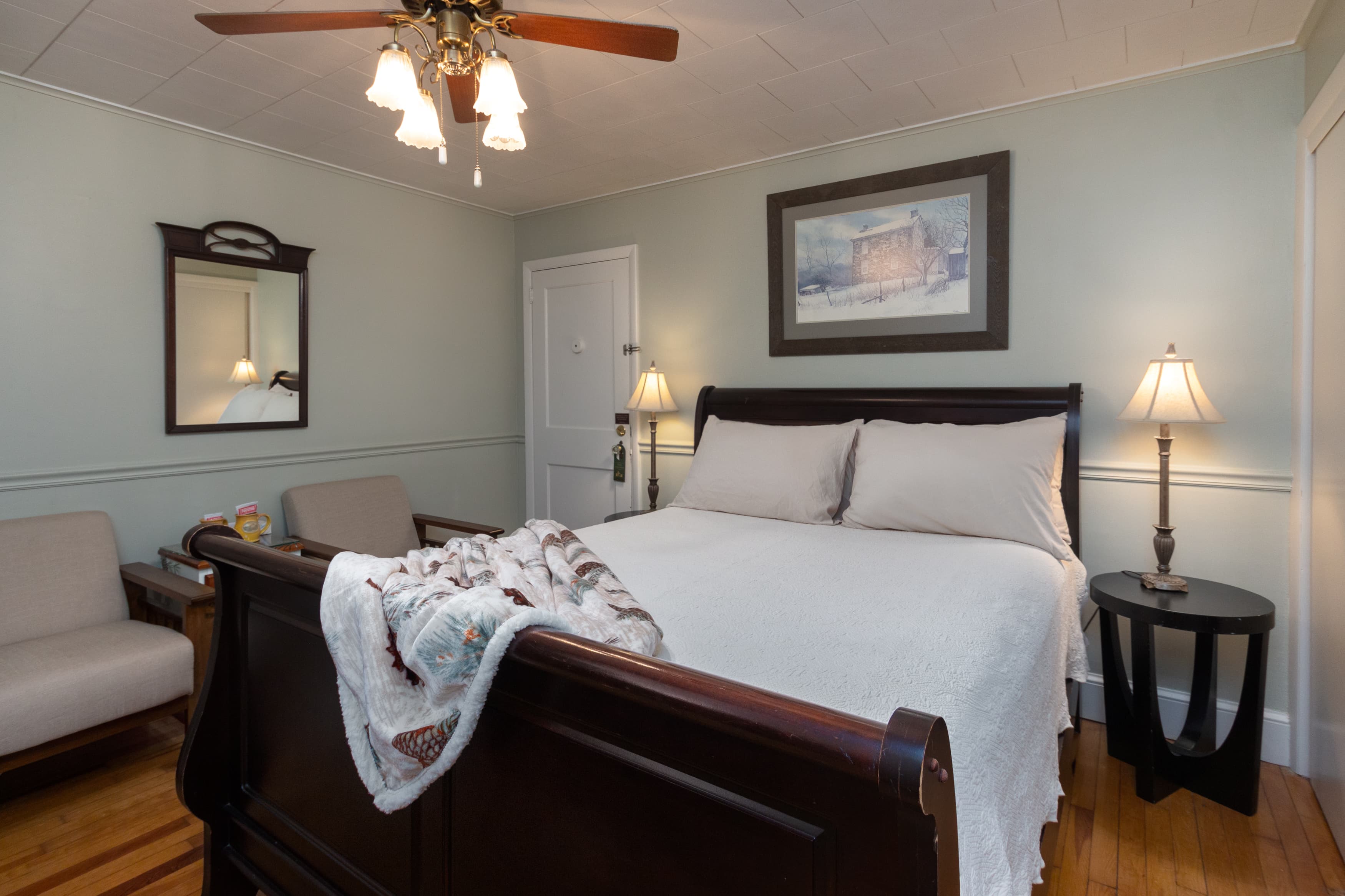 A tranquil bedroom with light green walls, a dark wood sleigh bed with a white duvet, and a small sitting area with two armchairs.