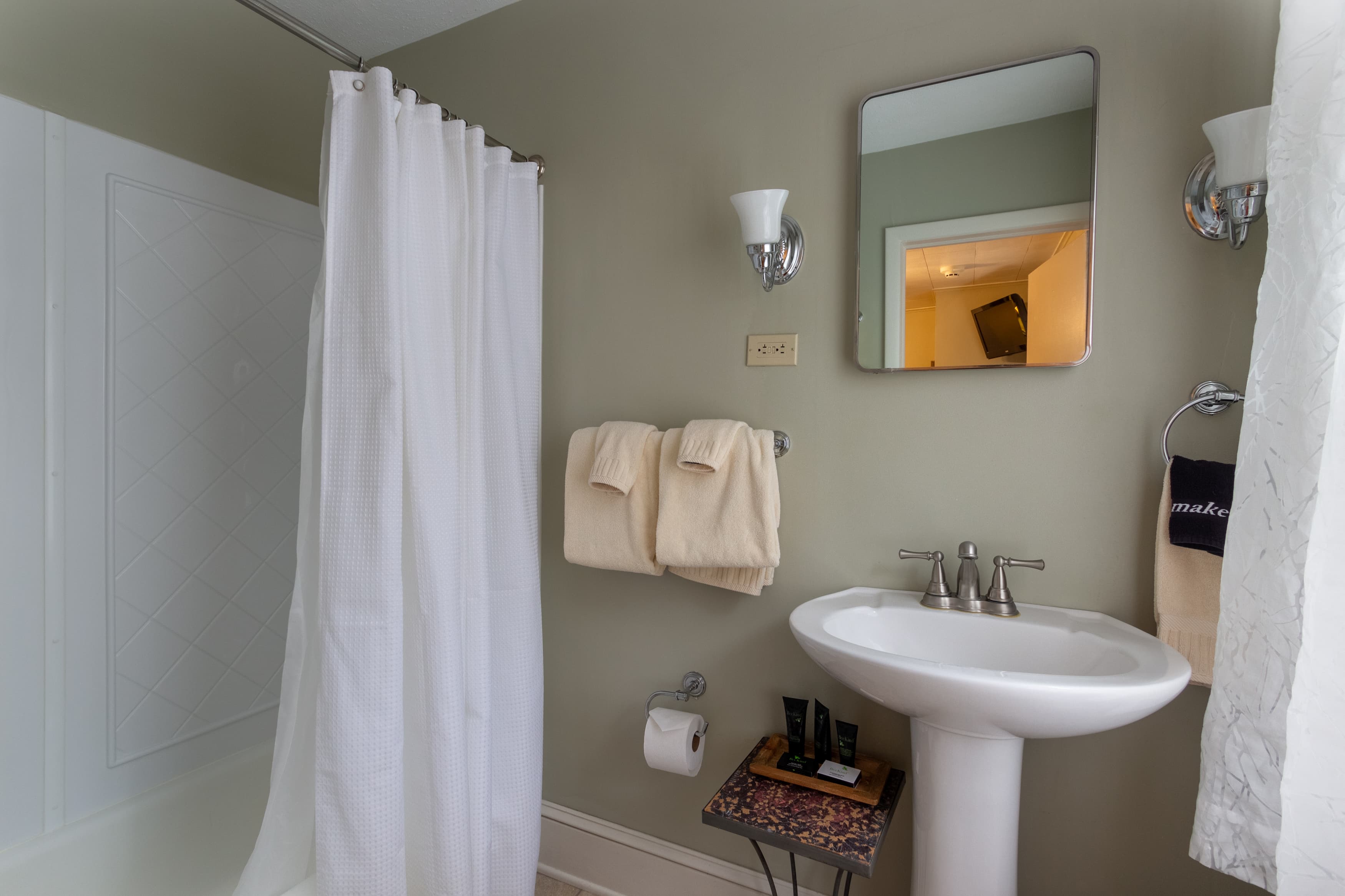 A clean, contemporary bathroom with sage-green walls, a white pedestal sink, a mirror, and a shower/tub with a white curtain.