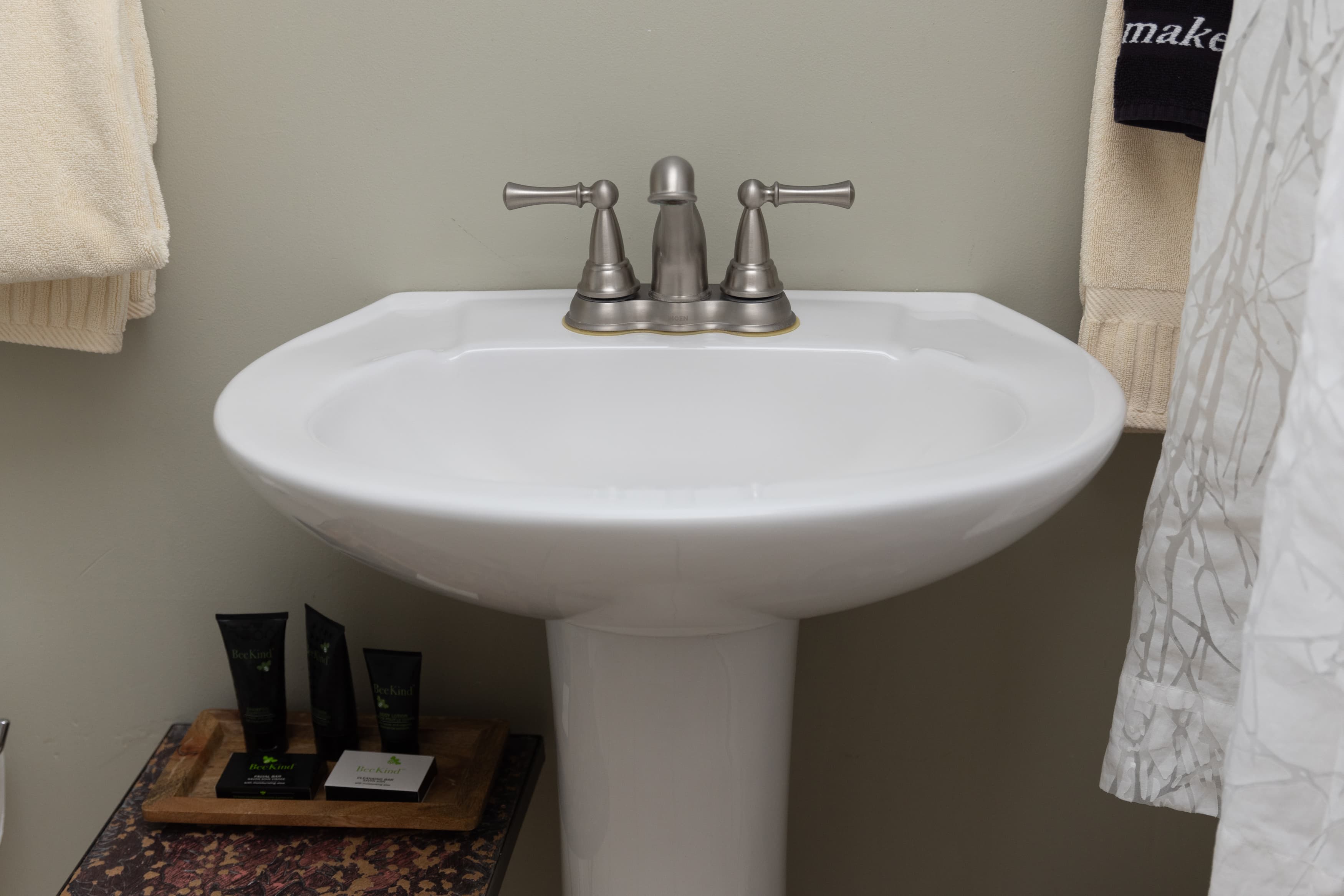A close-up of a white pedestal sink with brushed nickel faucet handles and a small wooden tray of toiletries at the base.