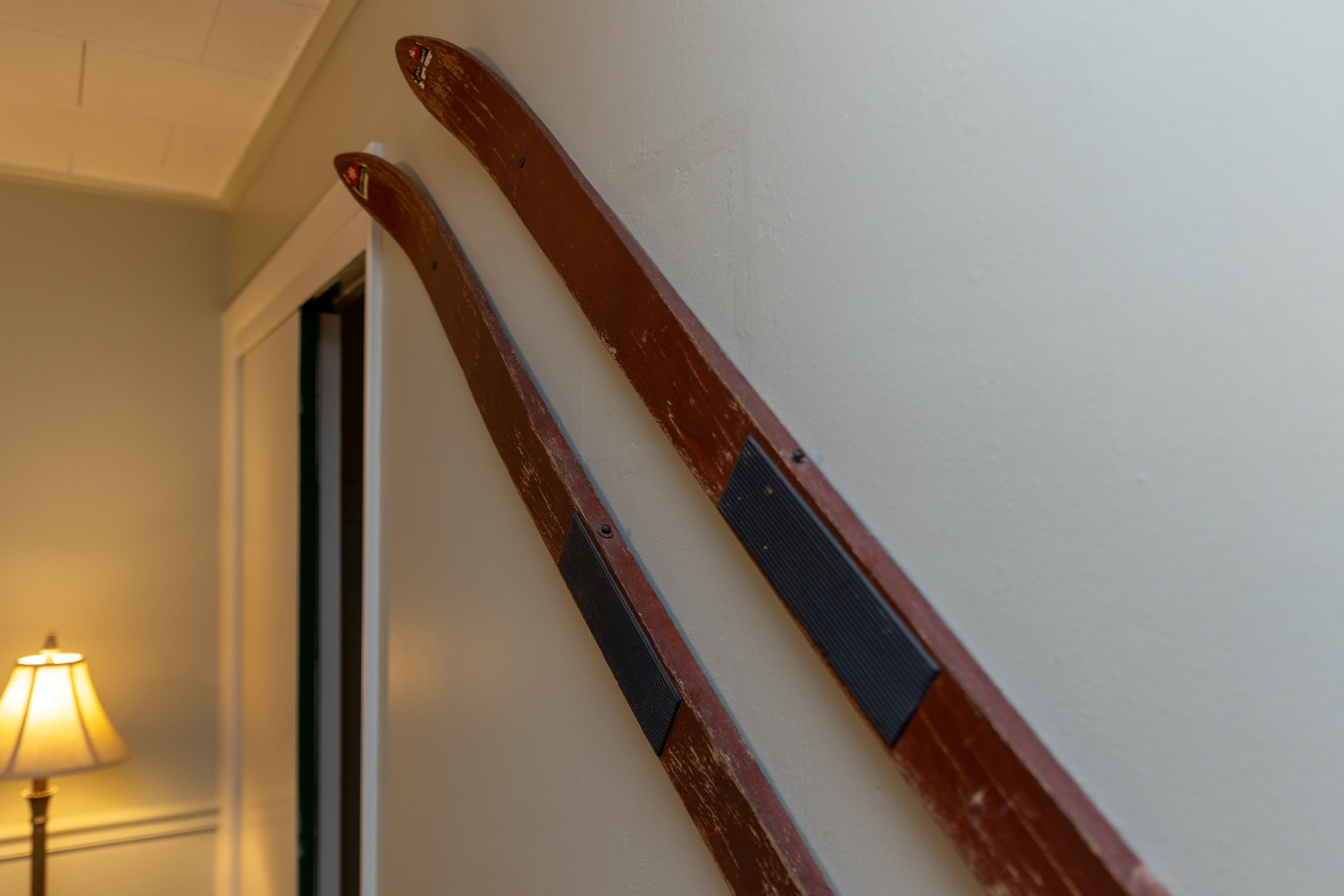 A close-up of a pair of vintage wooden skis with red-stained tips mounted diagonally on a light beige wall next to a doorway.