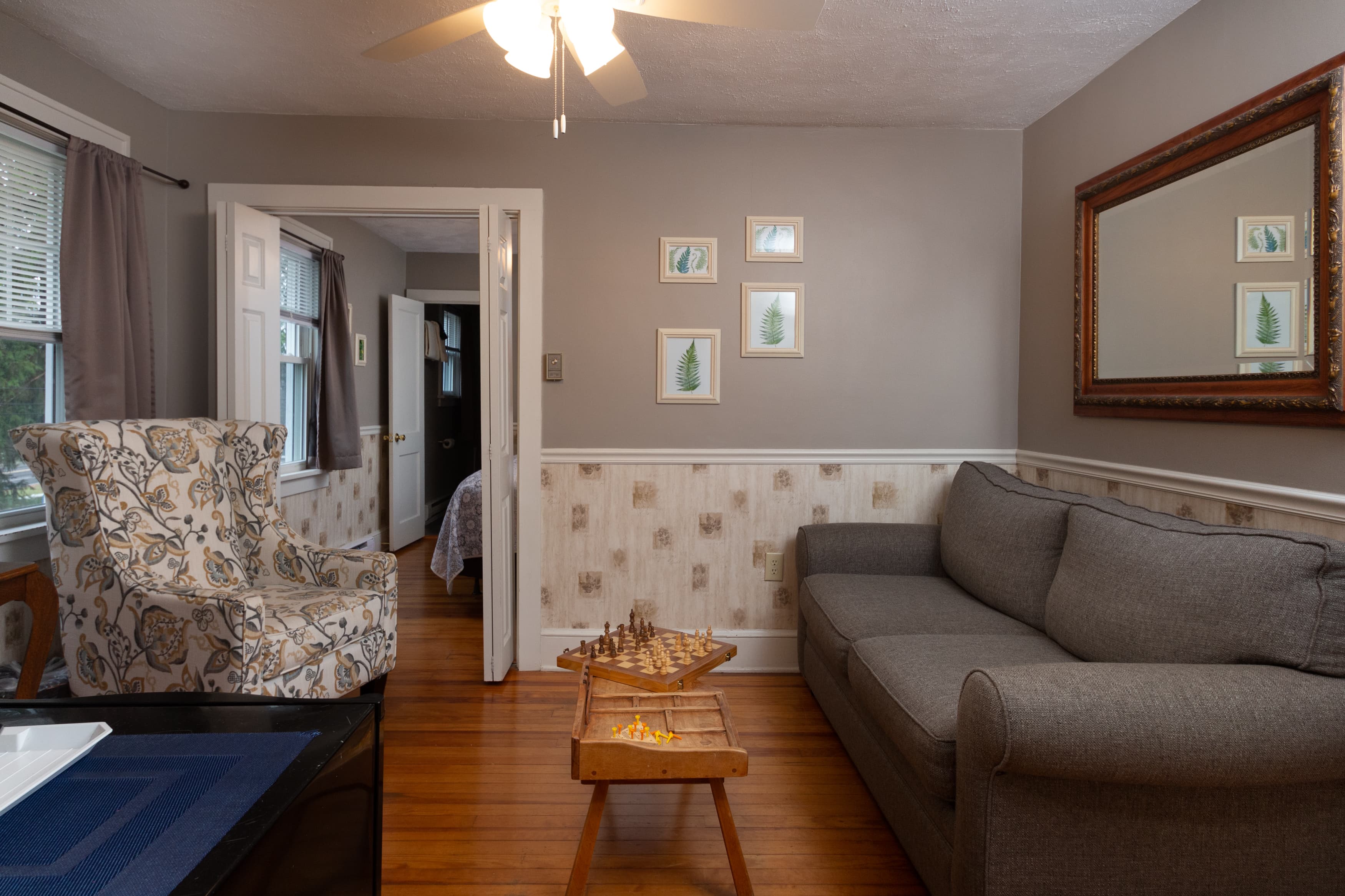 A suite living room with a gray sofa, a patterned armchair, hardwood floors, and a large framed mirror over a paneled wall.