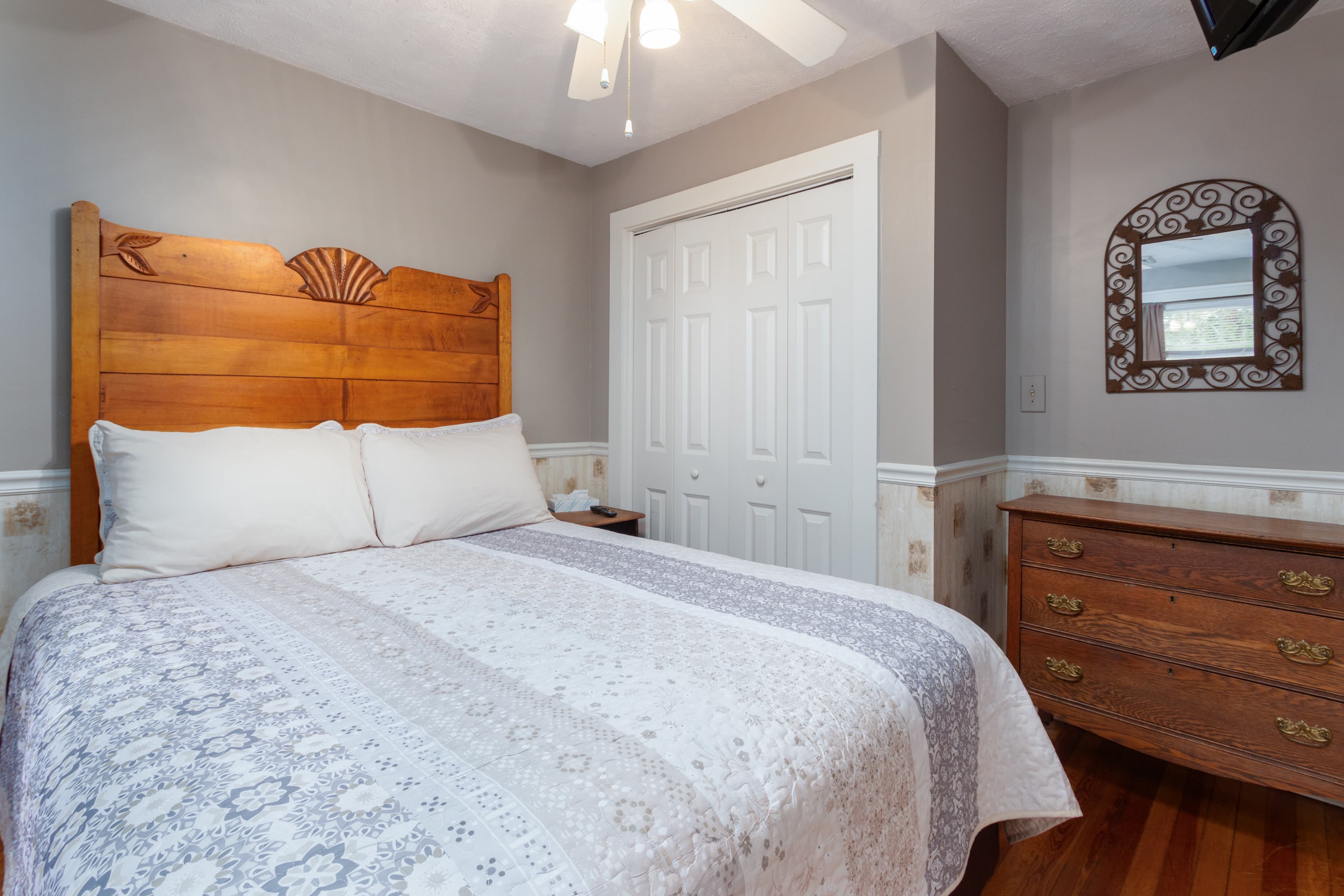A cozy bedroom with a wooden headboard, a white and blue patterned quilt, a dark wood dresser, and a closet with bi-fold doors.