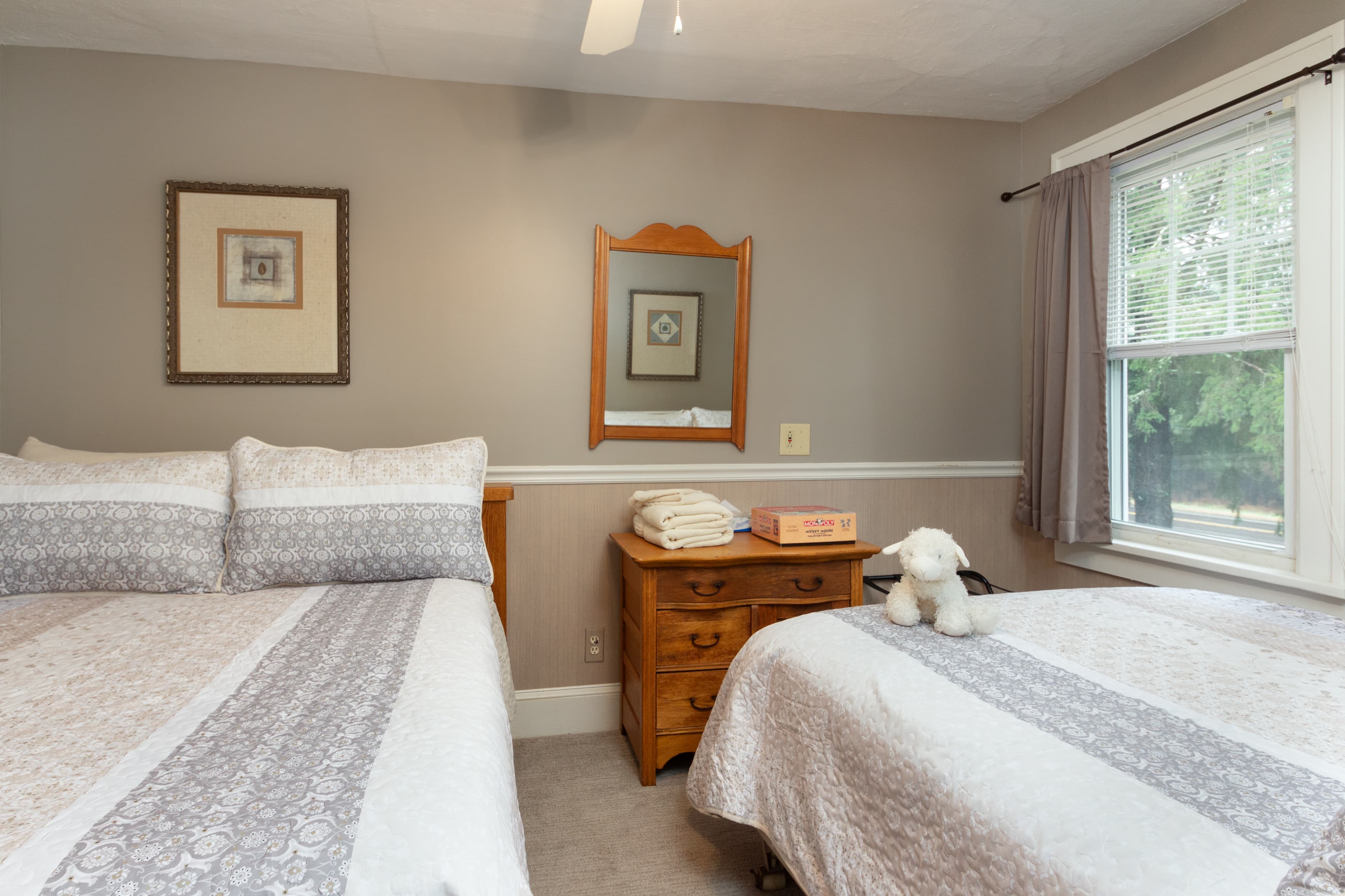 A cozy guest room with two full beds featuring patterned quilts, a light wood dresser with a mirror, and neutral gray walls.