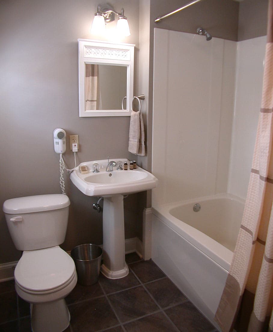 A clean, modern bathroom with gray walls, a white pedestal sink, a toilet, a tub/shower combo, and dark brown floor tiles.