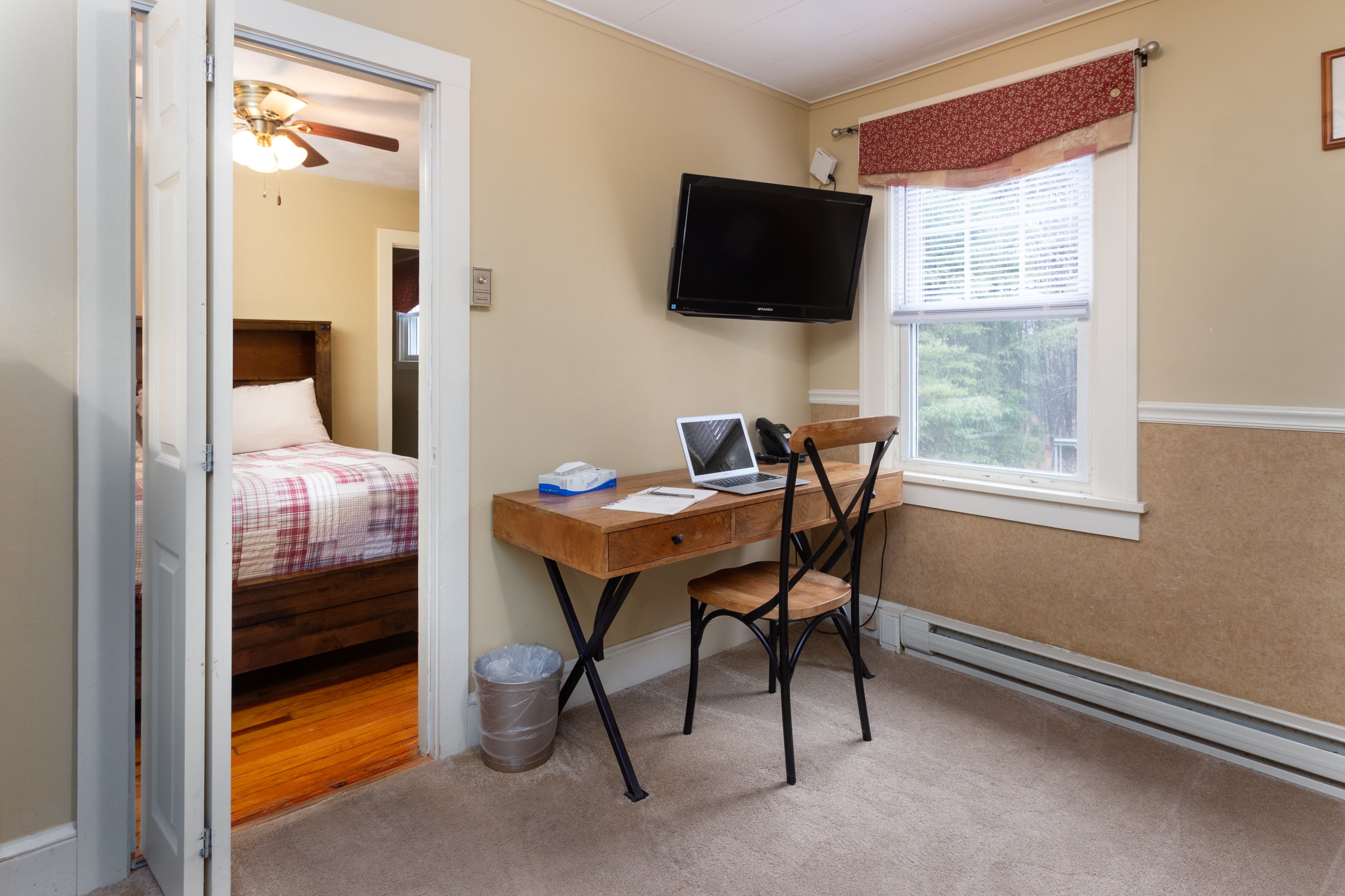 A small office nook with a rustic wood desk, a wall-mounted TV above a window, and a view through a doorway into the bedroom.