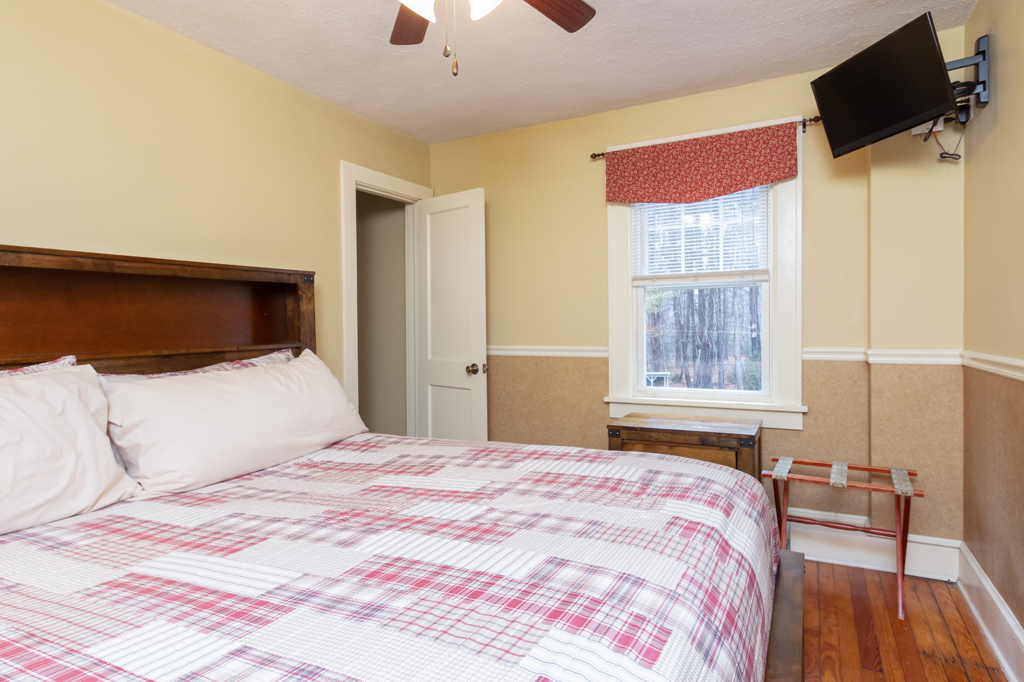 A cozy bedroom with a dark wood bed, a red and white patchwork quilt, a wall-mounted TV, and a storage chest near the window.