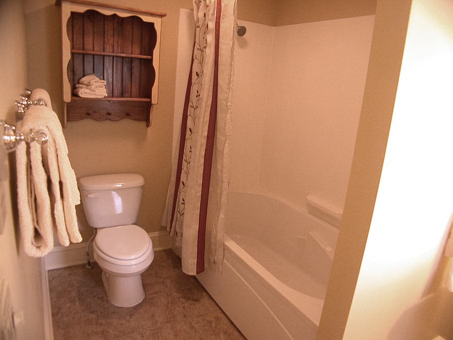A bathroom with a white toilet next to a bathtub/shower combo, featuring a wood shelf and a towel rack on the beige wall.