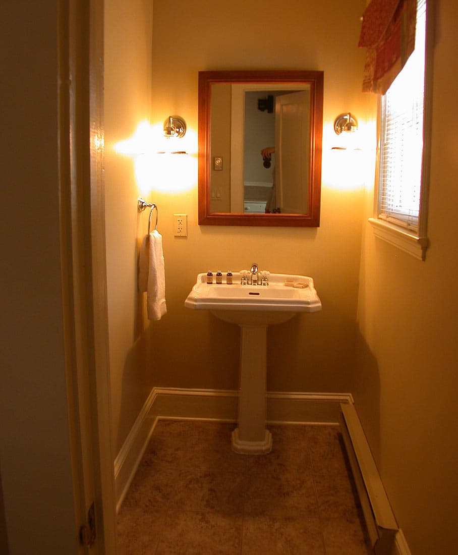 A small, brightly lit vanity area with a white pedestal sink, a wood-framed mirror, and sconce lights on warm yellow walls.