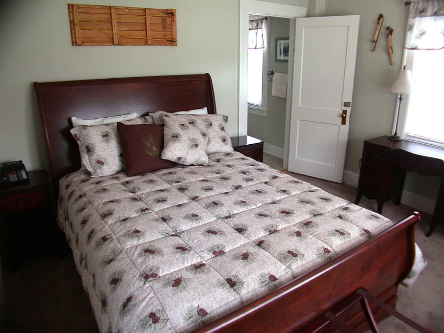 A bedroom with a dark wood sleigh bed and a floral quilt, featuring a desk by the window and a doorway leading to the bathroom.
