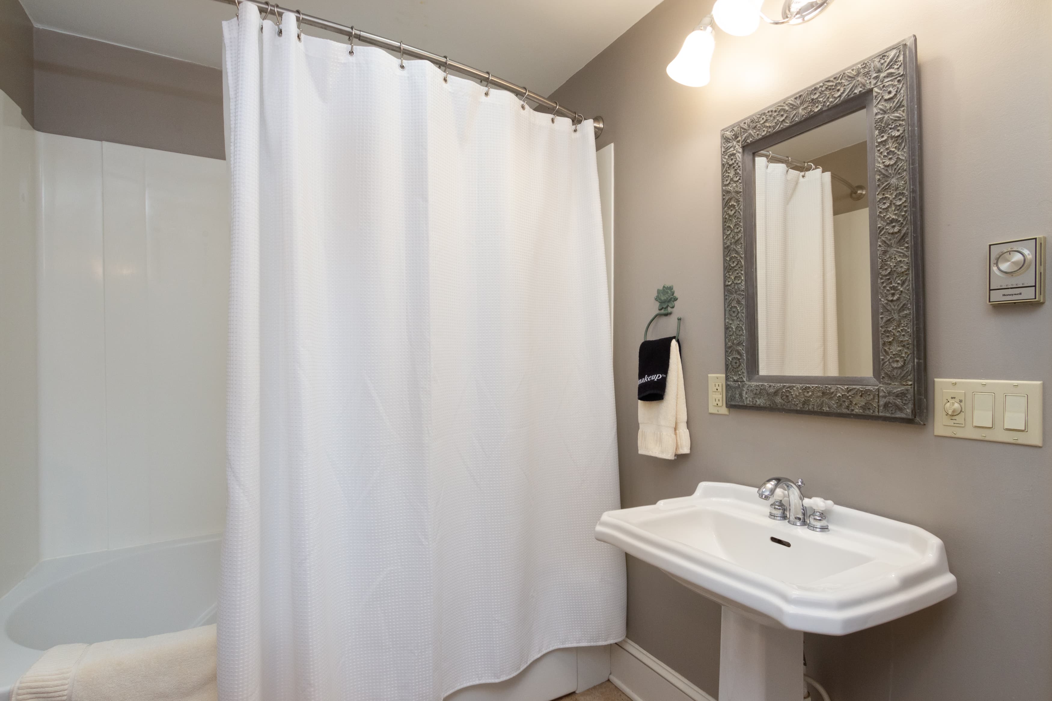 A clean, contemporary bathroom with a white pedestal sink, a decorative mirror, a tub/shower combo, and light taupe walls.