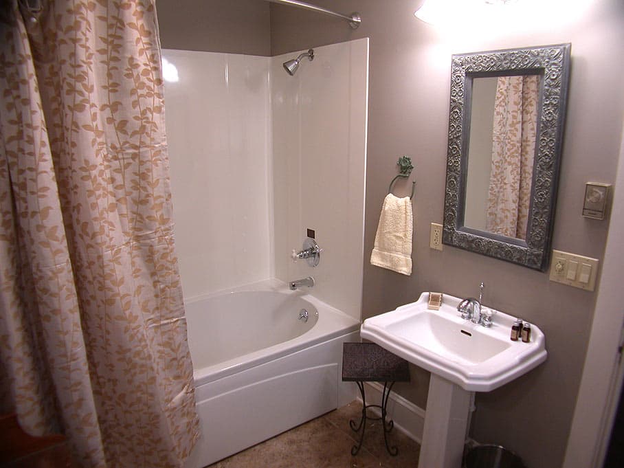 A clean, contemporary bathroom with a white pedestal sink, a gray-framed mirror, a tub/shower combo, and a patterned curtain.
