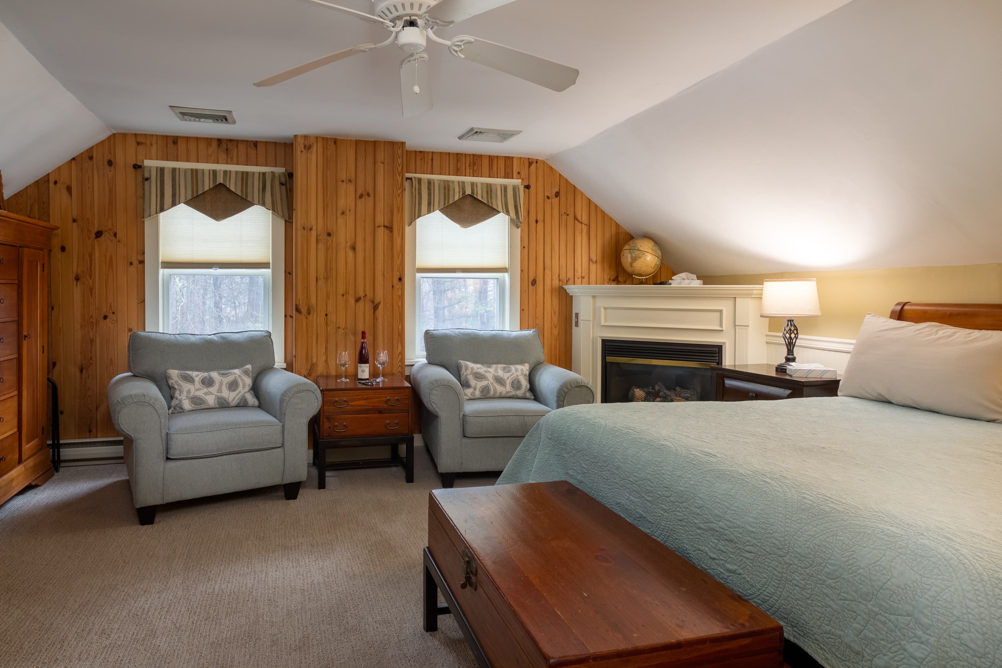 A third-floor bedroom with sloping ceilings, a wood-paneled wall behind two light blue armchairs, a king bed, and a fireplace.