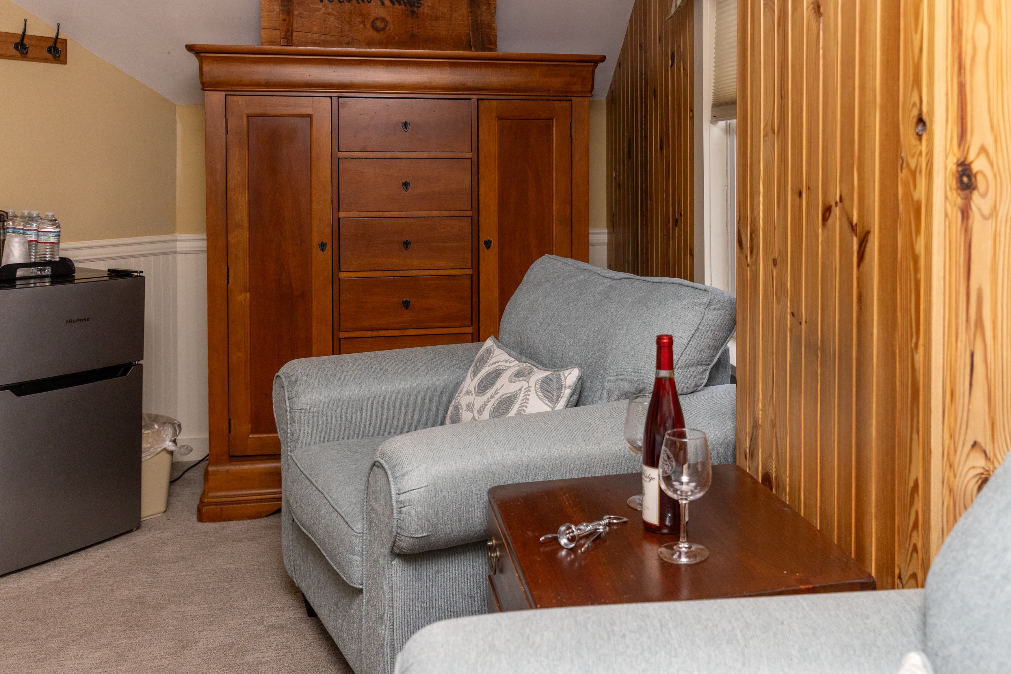 A sitting area with a light blue armchair, a dark wood armoire/dresser, a mini-fridge, and a wine bottle on a side table.