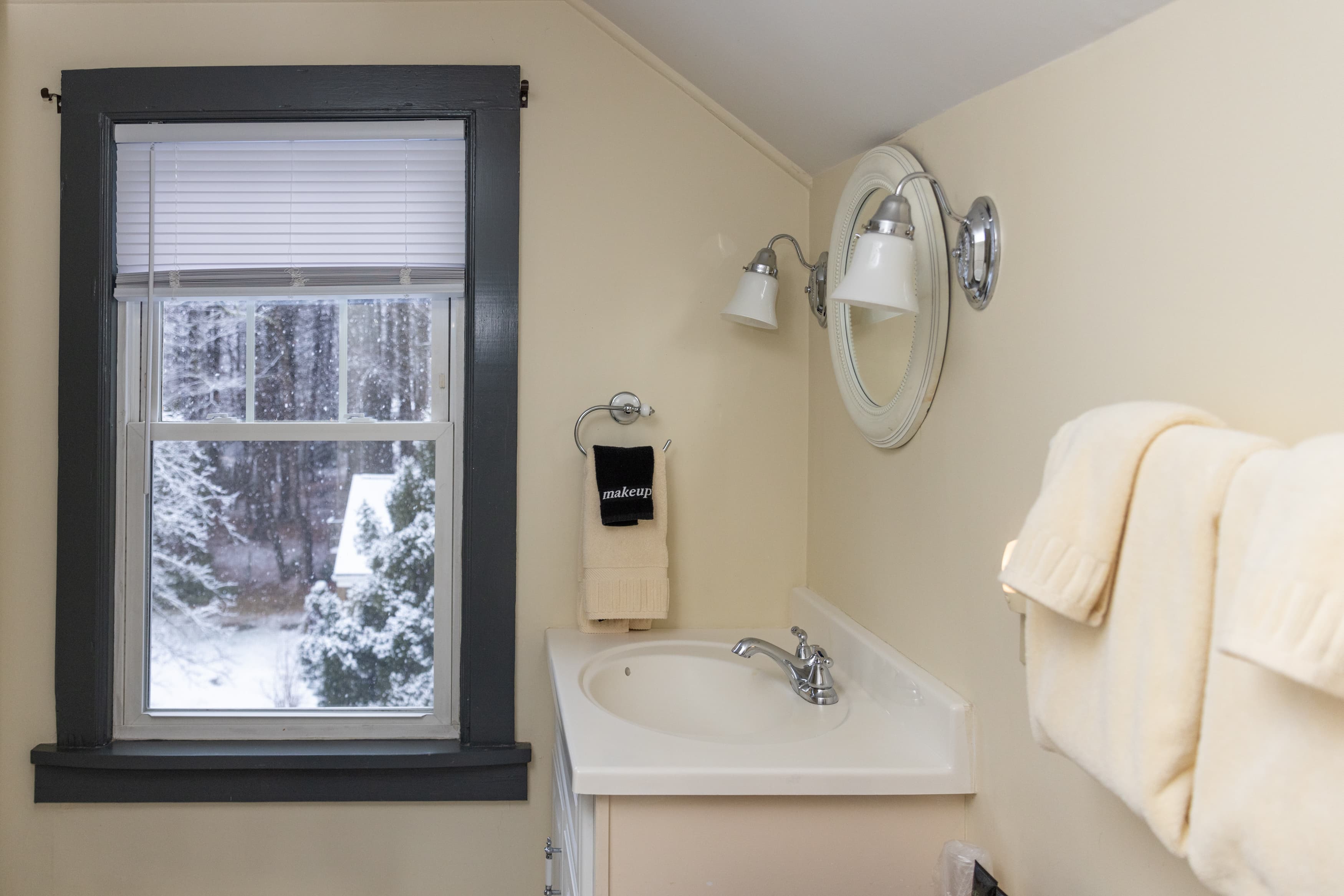 A bright bathroom vanity area with a window view of snowy trees, a sink in a light vanity, an oval mirror, and light yellow walls.