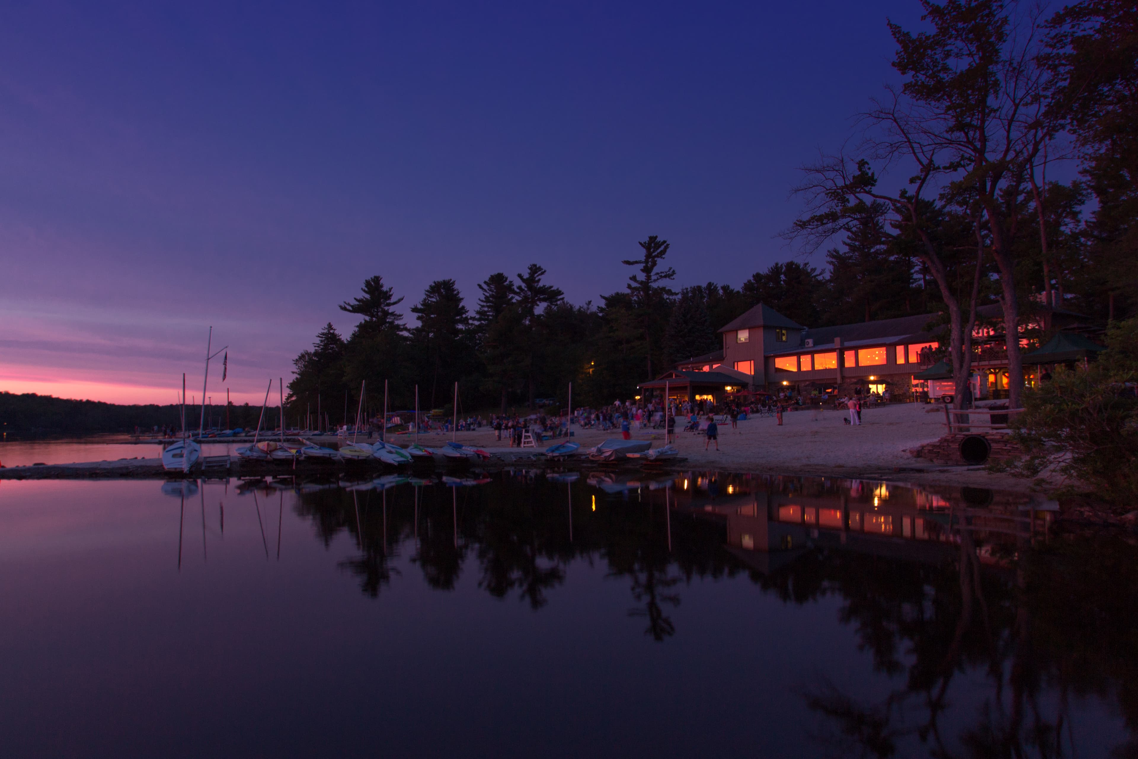 A tranquil lakefront scene at dusk with illuminated buildings and anchored boats reflecting on the water.