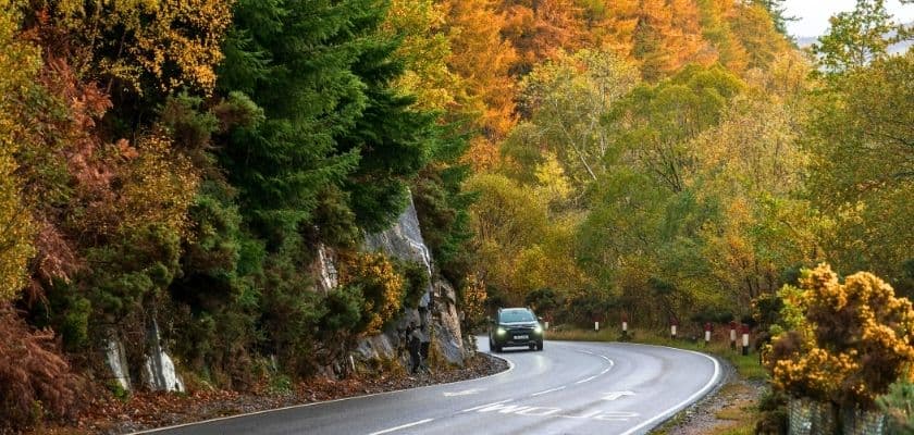 A car drives along a winding road surrounded by autumn-colored trees.