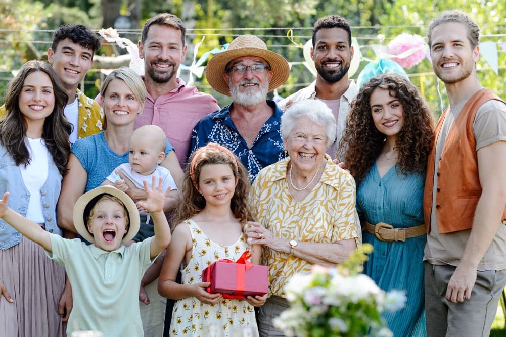 A joyful multigenerational family gathers outdoors, smiling together while holding a small gift.