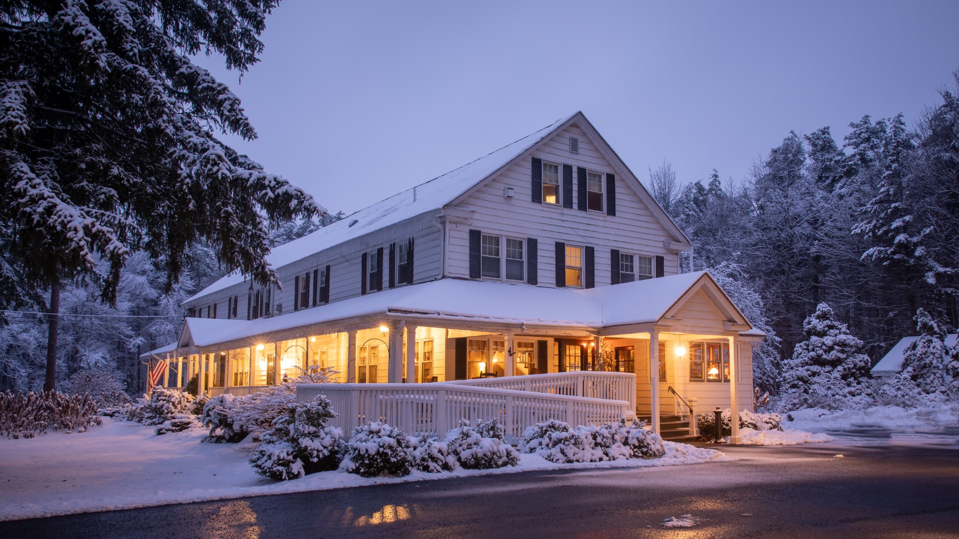 A cozy white house illuminated at dusk, surrounded by fresh snow and frosty trees.