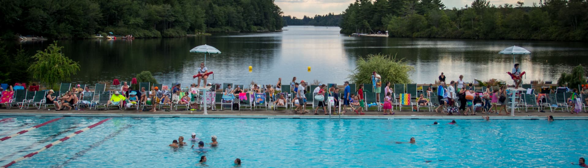 A crowded outdoor pool area is filled with people lounging on chairs and swimming, overlooking a serene lake.