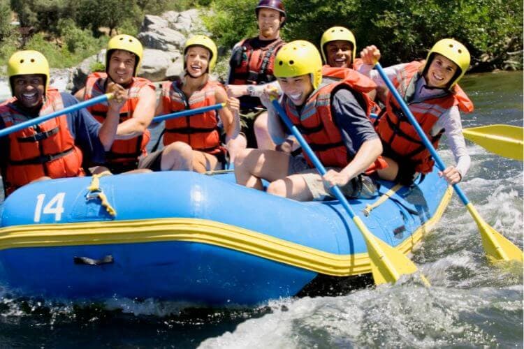 A group of six people joyfully paddles a blue raft on a river.