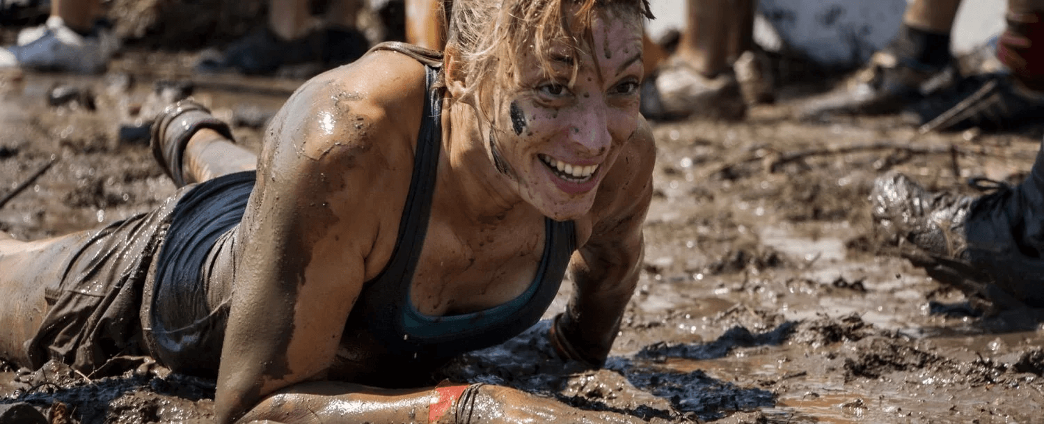 A muddy participant smiles while crawling through a dirt-covered obstacle course.