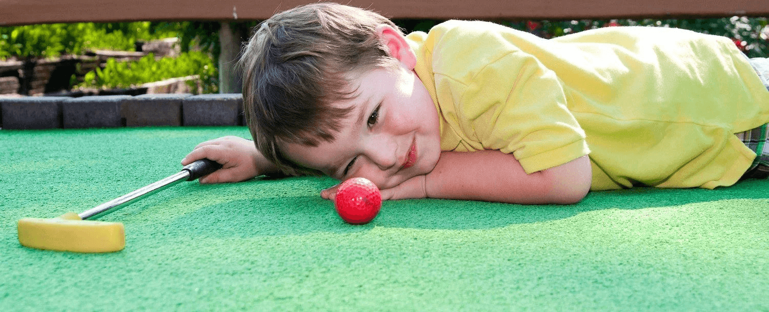 A young boy in a yellow shirt lies on a green mini-golf course, closely examining a red ball with a putter nearby.