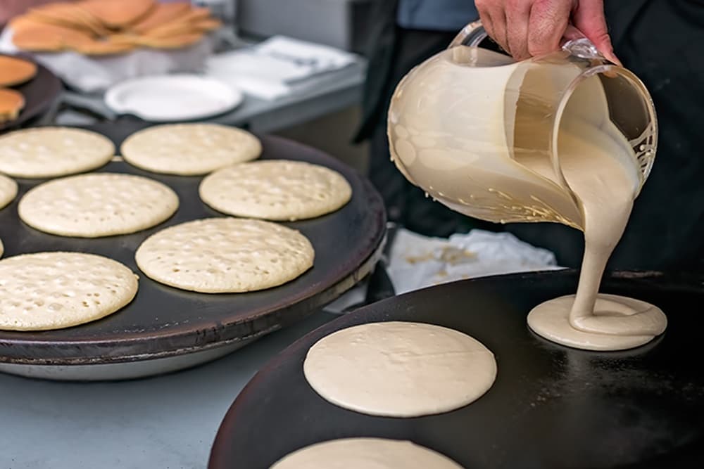 A person pours pancake batter onto a hot griddle with several pancakes cooking nearby.