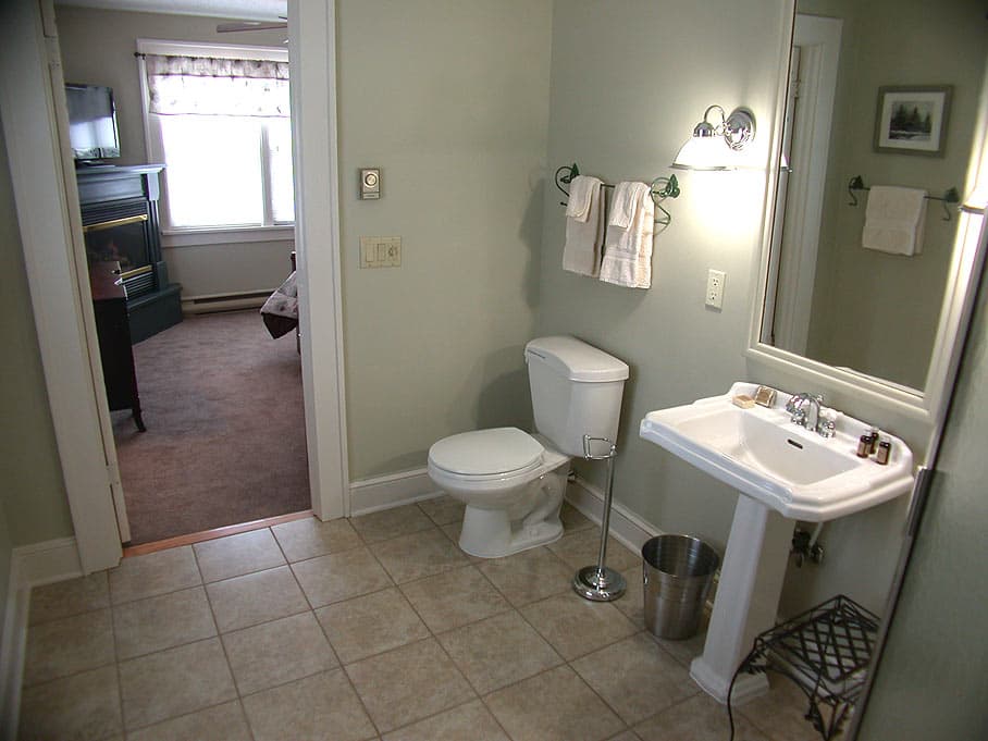 A simple, spacious bathroom with sage-green walls, a white pedestal sink and toilet, with a view into the adjacent bedroom.