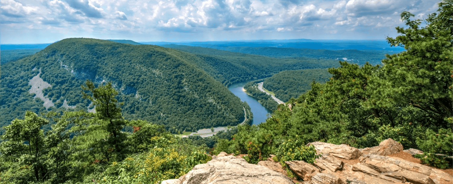 A panoramic view of a lush valley with a winding river and rolling hills under a partly cloudy sky.