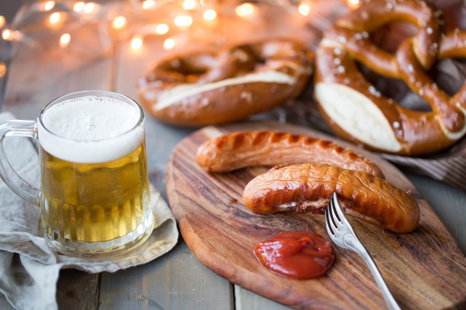 A wooden board with a sausage, fork, ketchup, pretzels, and a mug of beer, set against a backdrop of warm lights.