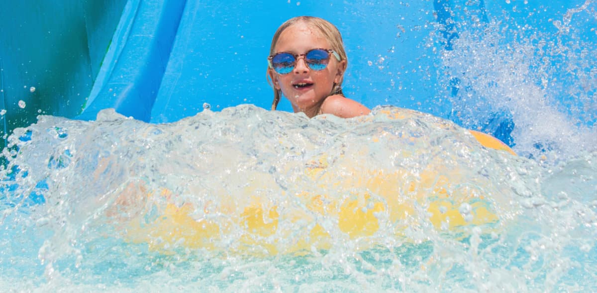 A child on a yellow inflatable tube splashes through water at a water slide.