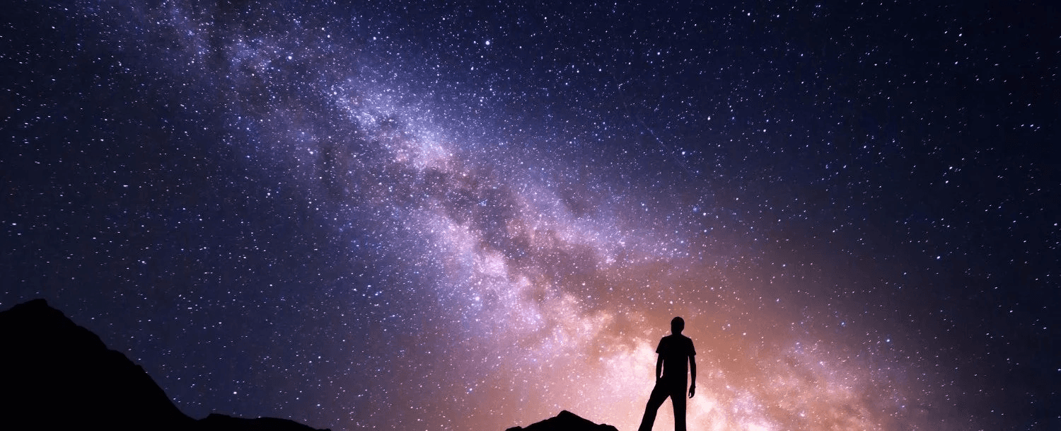 Silhouette of a person standing on a rocky outcrop under a star-filled sky with the Milky Way.