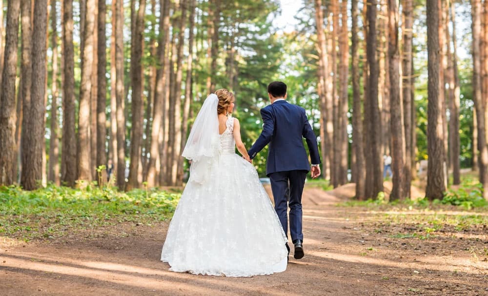 A couple walks hand in hand down a woodland path, dressed in wedding attire.