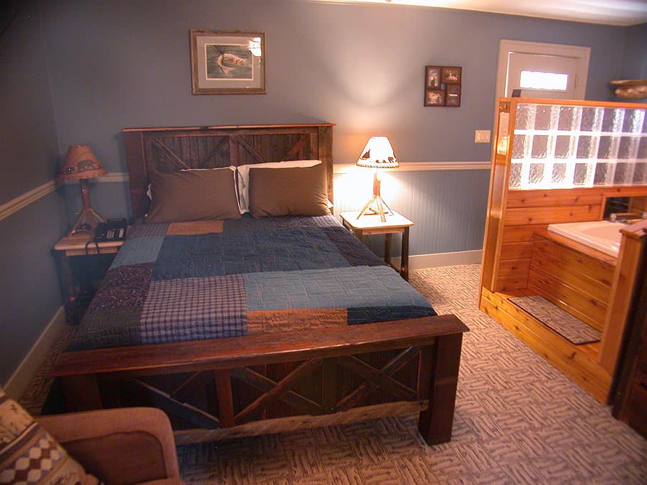 A rustic-themed hotel room with a dark wood bed frame, a blue and brown quilt, and a jetted tub surrounded by wood and glass blocks.