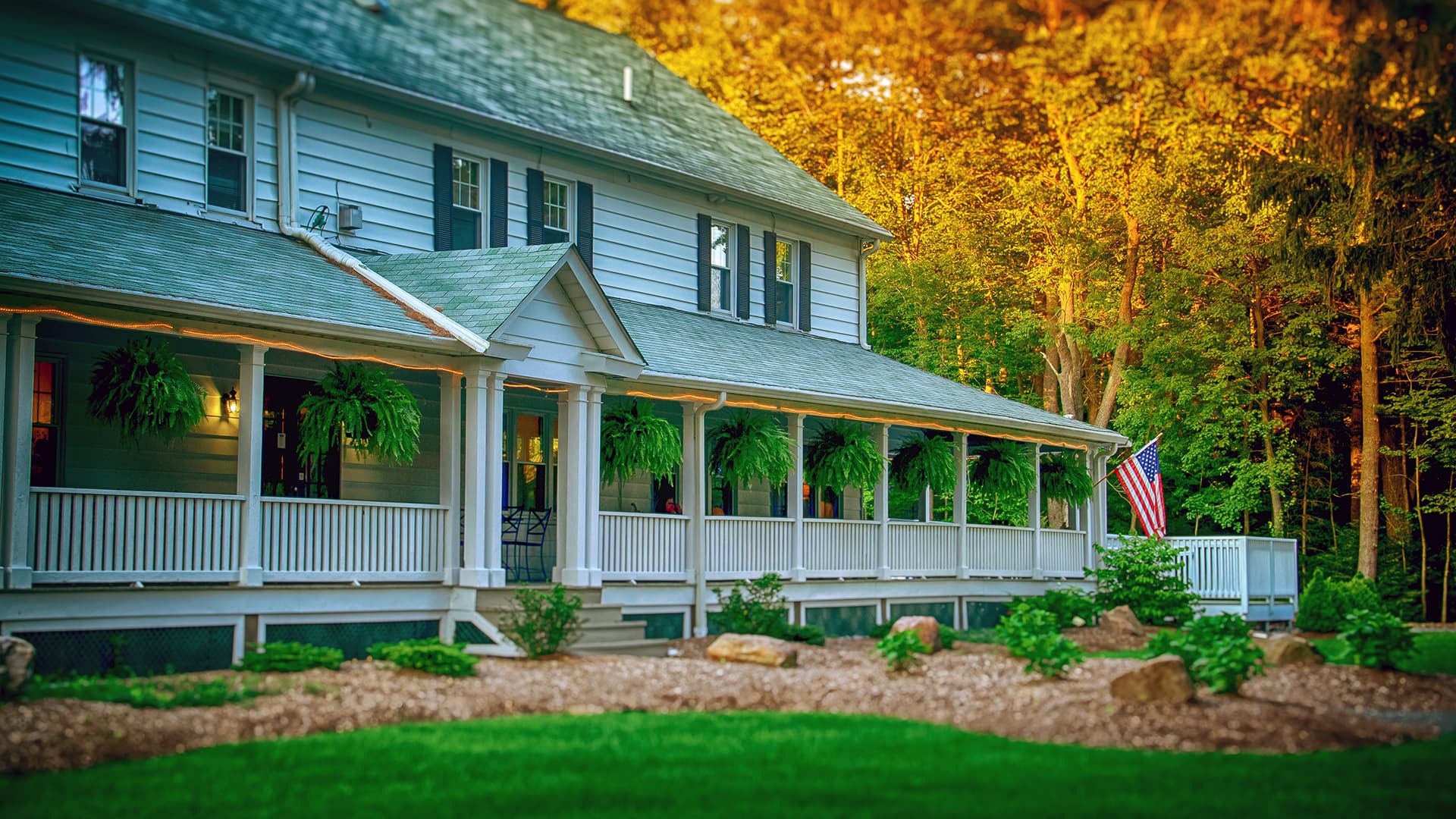 A charming two-story house with a front porch adorned with ferns and an American flag, surrounded by lush greenery.