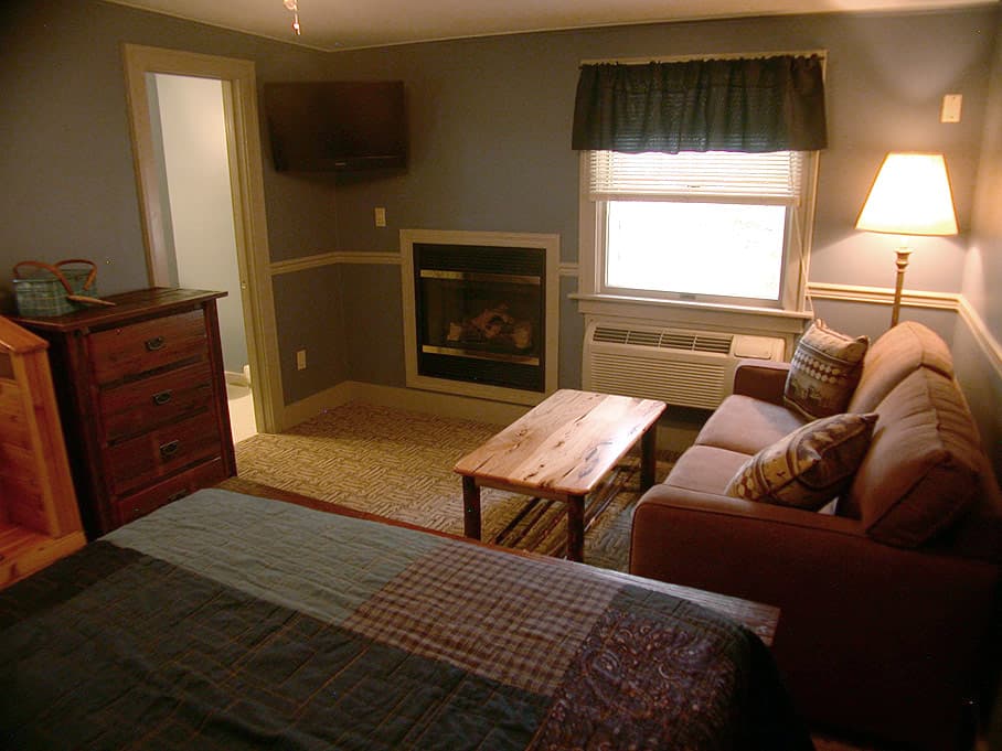 A cozy hotel room with a queen bed, a dark-colored sofa, a rustic coffee table, and a built-in fireplace on the blue wall.