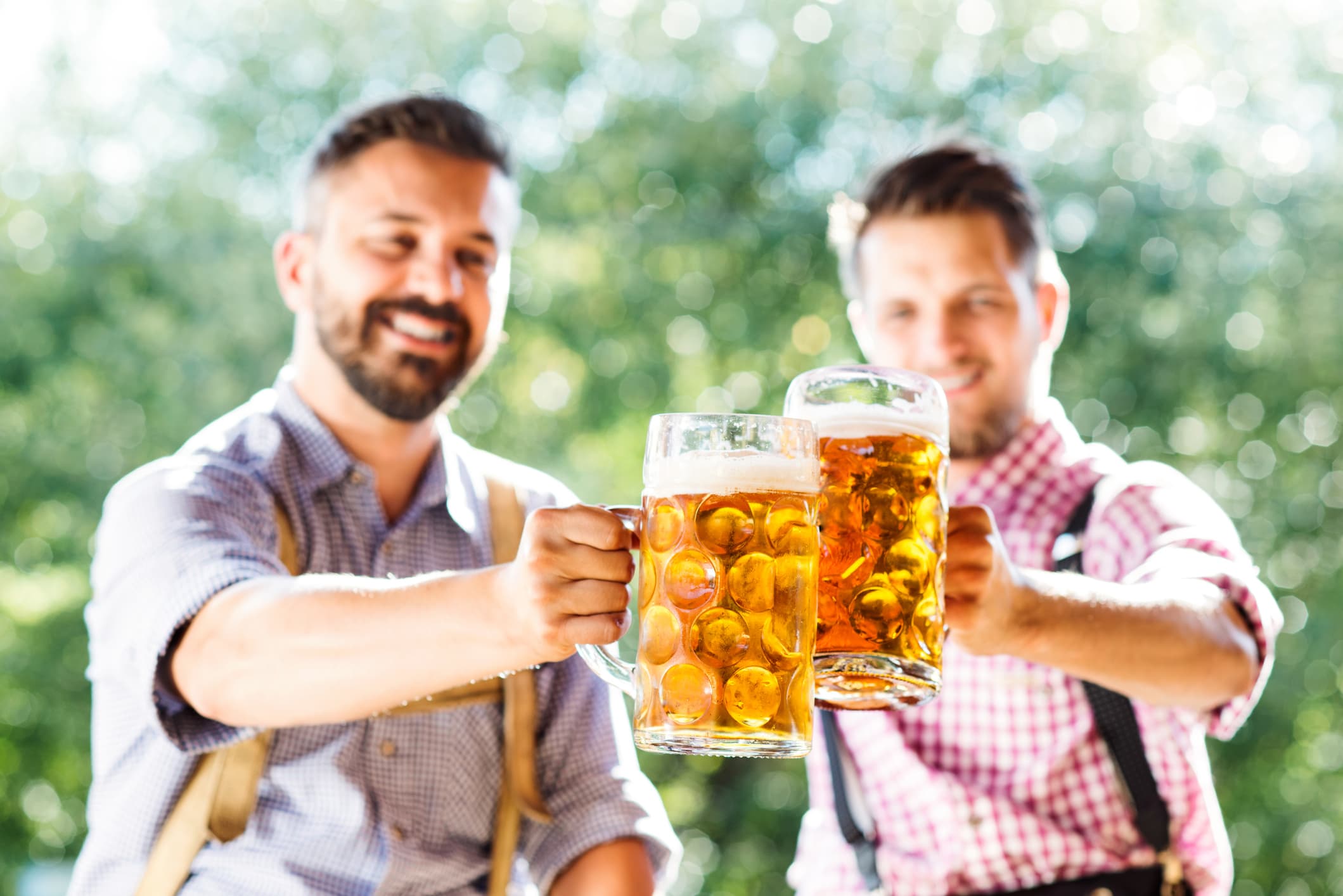 Two men clink large mugs of beer outdoors with a blurred background.