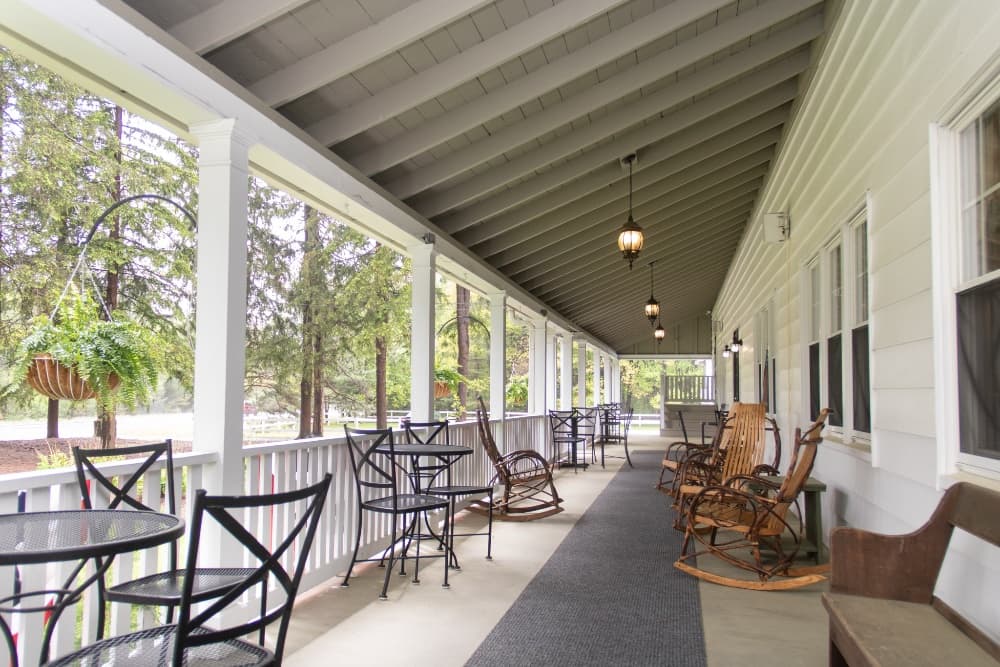 A covered porch featuring rocking chairs, tables, and hanging plants, surrounded by trees.