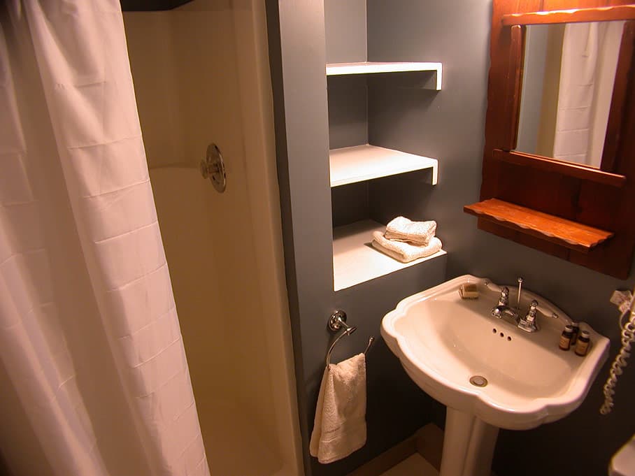 A compact bathroom featuring a white pedestal sink, built-in shelving with towels, a wooden mirror, and a shower curtain.
