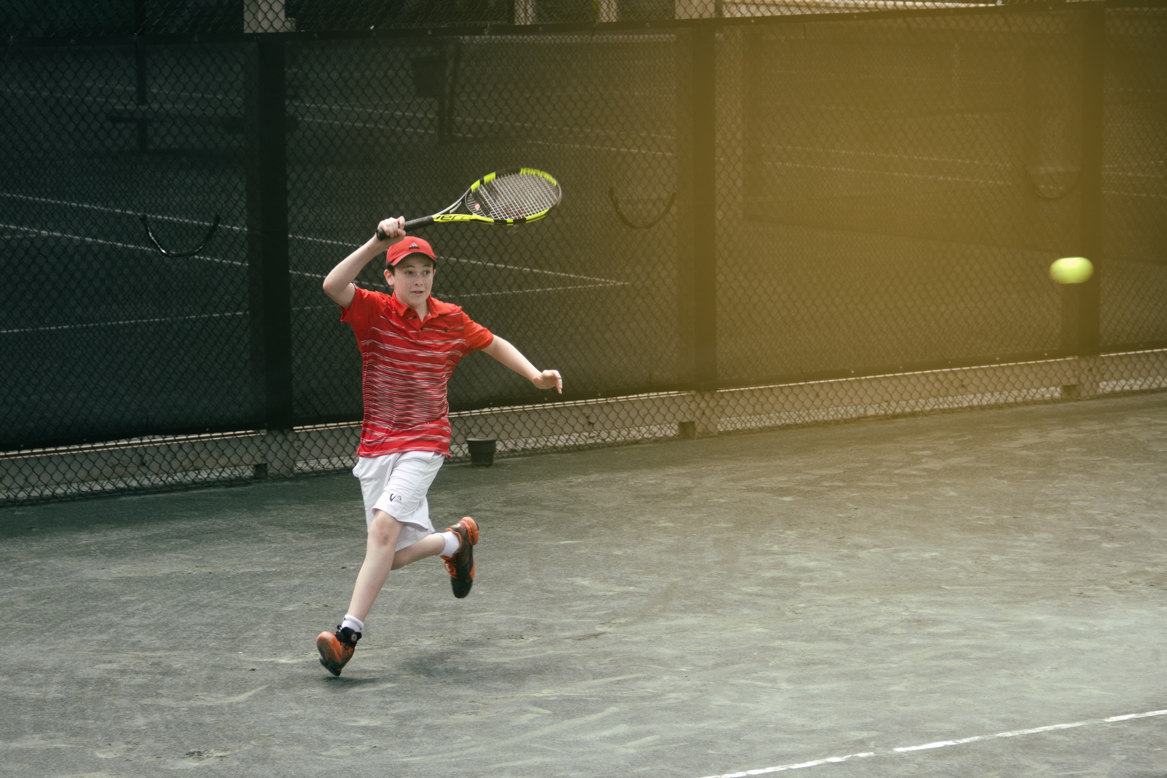 A young boy in a red striped shirt and cap is playing tennis on a court.
